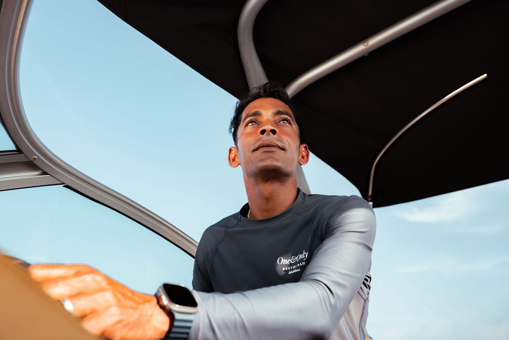 Adult man in his 30s with short dark hair steers a boat, wearing a fitted long-sleeve top, looking ahead under a shaded canopy against a clear blue sky.