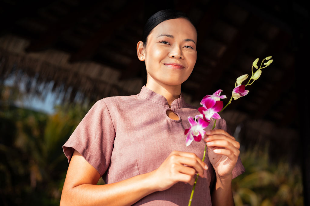 Adult woman in her 20s or 30s with dark hair tied back smiles gently while holding a pink orchid stem outdoors, wearing a mauve spa uniform beneath warm sunlight.