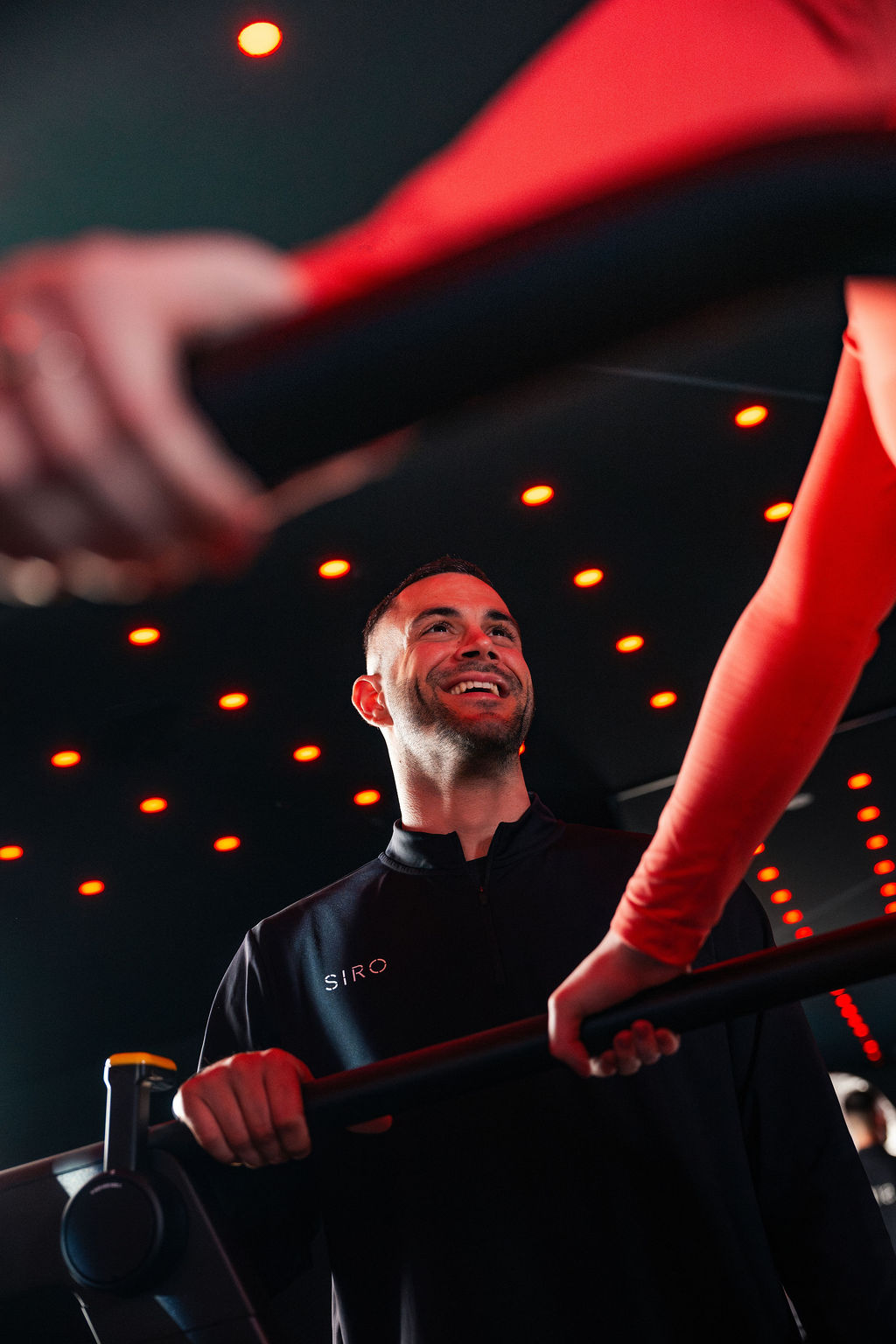 Adult man in his 20s or 30s with short dark hair smiles while coaching a workout participant, wearing a black SIRO jacket inside a red-lit training space.