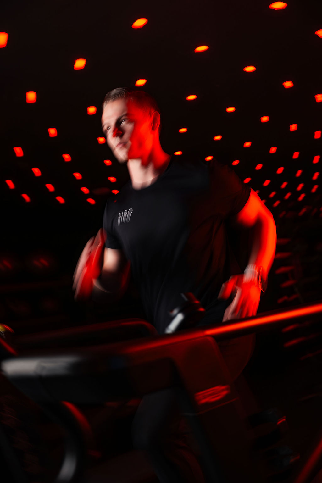 Adult man in his 20s or 30s with short light brown hair runs on a treadmill, wearing a black SIRO T-shirt under dramatic red lighting in a high-energy fitness studio.