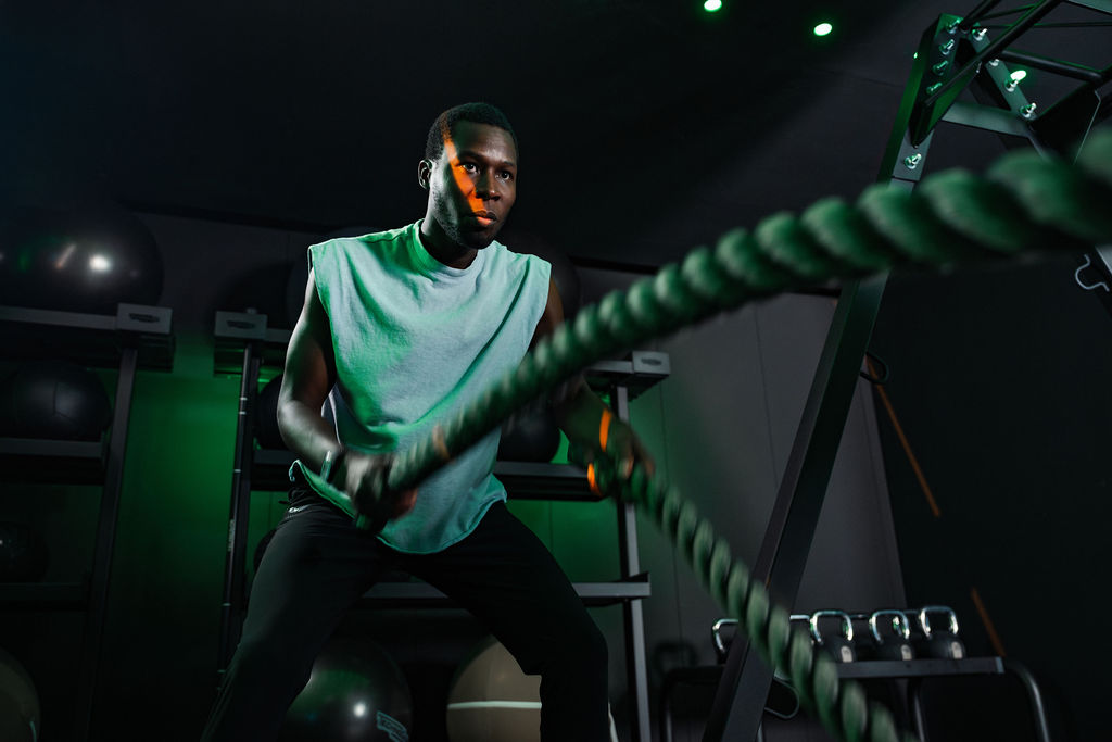 Adult man in his 20s or 30s with short dark hair performs battle rope exercises, wearing a sleeveless light top and black trousers inside a gym with green accent lighting.