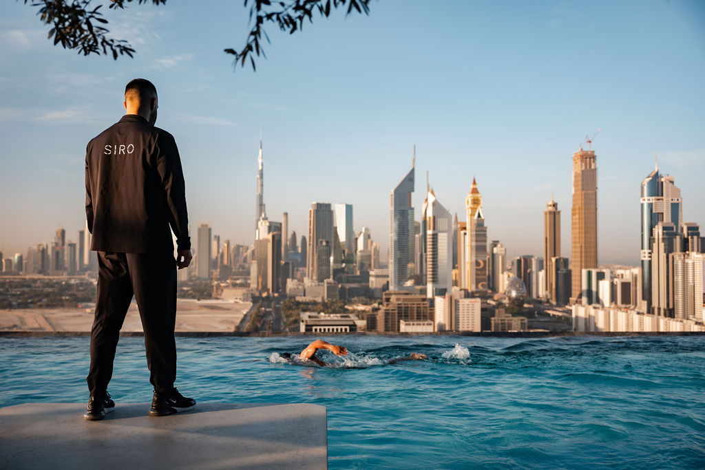 Adult man in his 20s or 30s with short dark hair stands poolside, wearing a black SIRO jacket, watching a swimmer in an infinity pool overlooking a modern city skyline.
