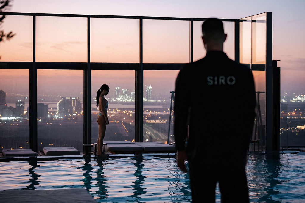 Adult man in his 20s or 30s stands beside a rooftop pool at sunset, wearing a black SIRO jacket, looking towards a woman standing near the water with city lights beyond.