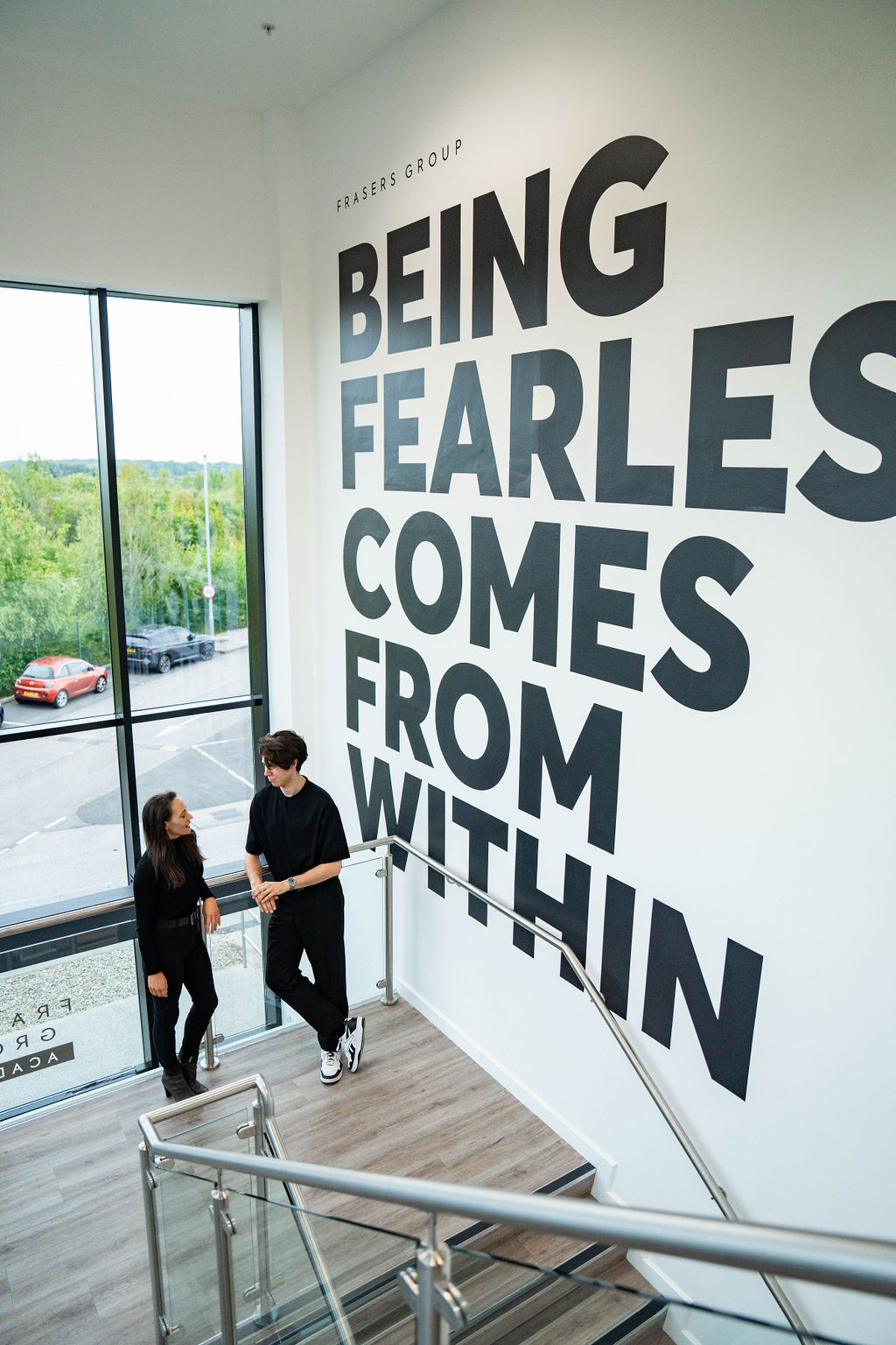 Two adults in their 20s or 30s stand on a stair landing beneath large wall text reading Being Fearless Comes From Within, talking casually in a bright office atrium.