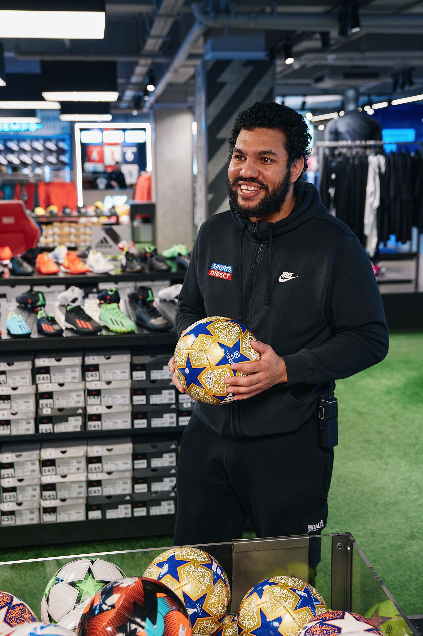 Sports Direct staff member smiling while holding a football in a modern store, surrounded by football boots and sportswear displays, creating a friendly, knowledgeable retail environment focused on customer service.