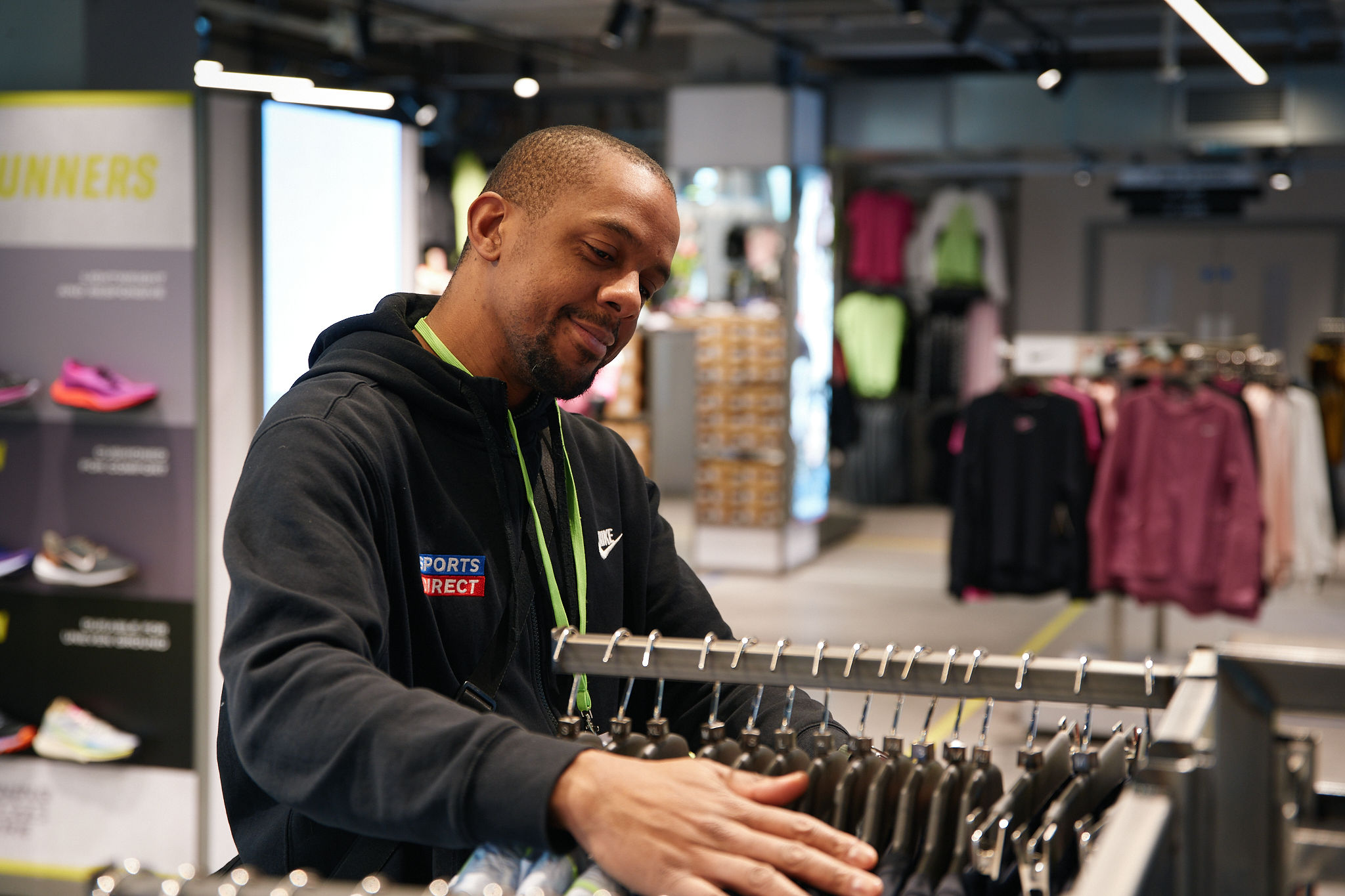 Sports Direct staff member organising clothing on a rail inside a spacious, well-lit store, wearing branded sportswear, with trainers and apparel displays visible, highlighting attentive retail care and product presentation.