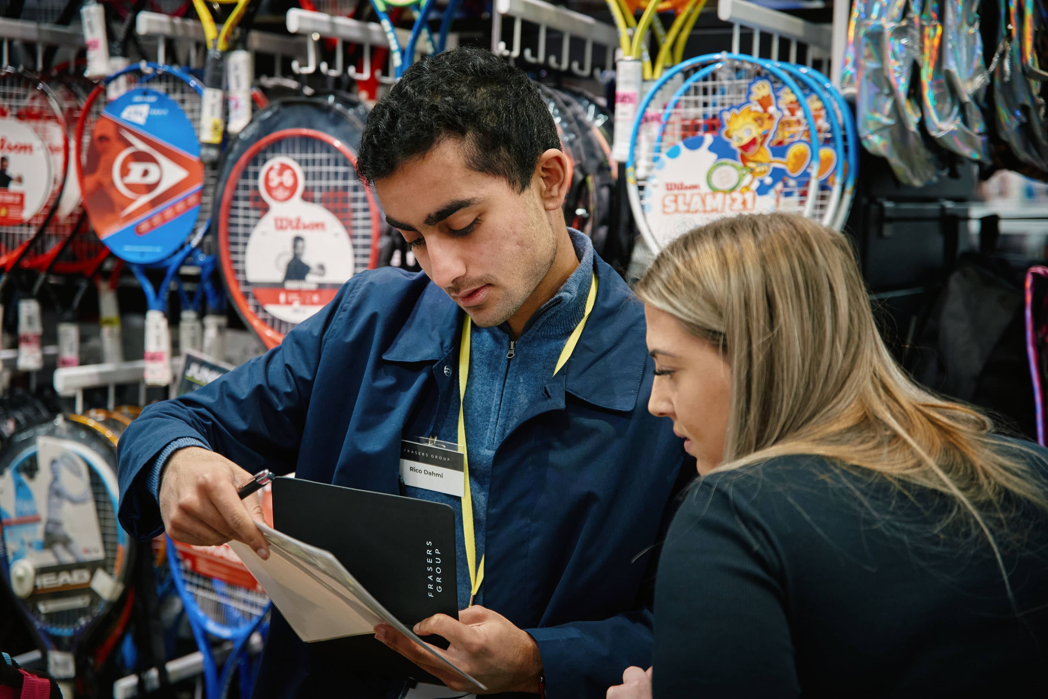 Man wearing a navy jacket and lanyard reviews documents with a woman in a black top inside a sports retail store, surrounded by tennis rackets and sporting equipment displays.
