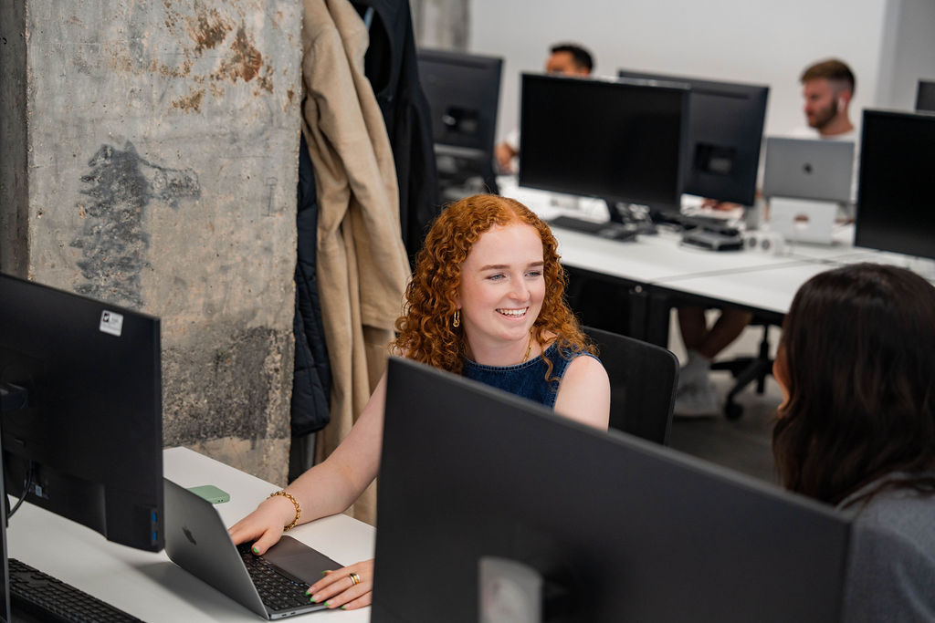 Woman with curly red hair wearing a sleeveless navy top smiles while working on a laptop at a desk, talking to a colleague in a modern open-plan office.