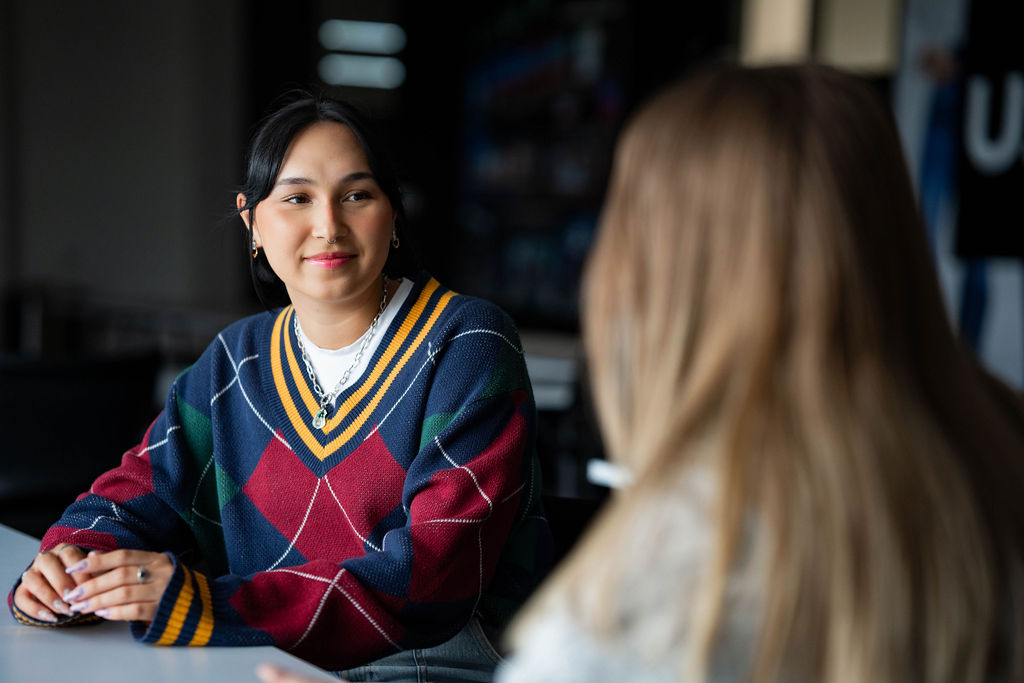 Woman wearing a patterned V-neck jumper sits at a table facing another woman, listening attentively during a conversation in a softly lit office or meeting area.