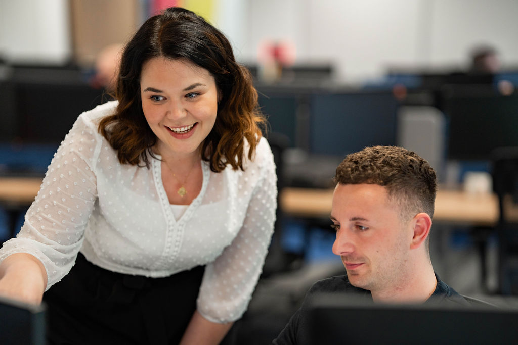 Woman wearing a white blouse gestures toward a computer screen while speaking to a seated man in a black T-shirt, collaborating at a desk in a shared office space.