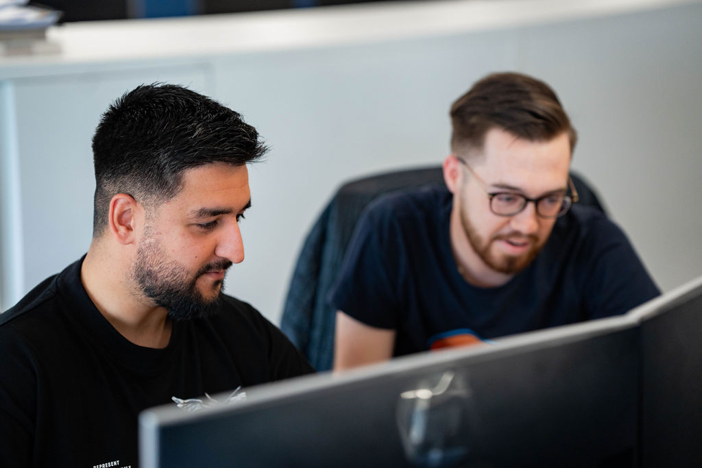 Two men wearing dark T-shirts sit side by side at desks, focused on computer screens, collaborating quietly in an open-plan office environment.