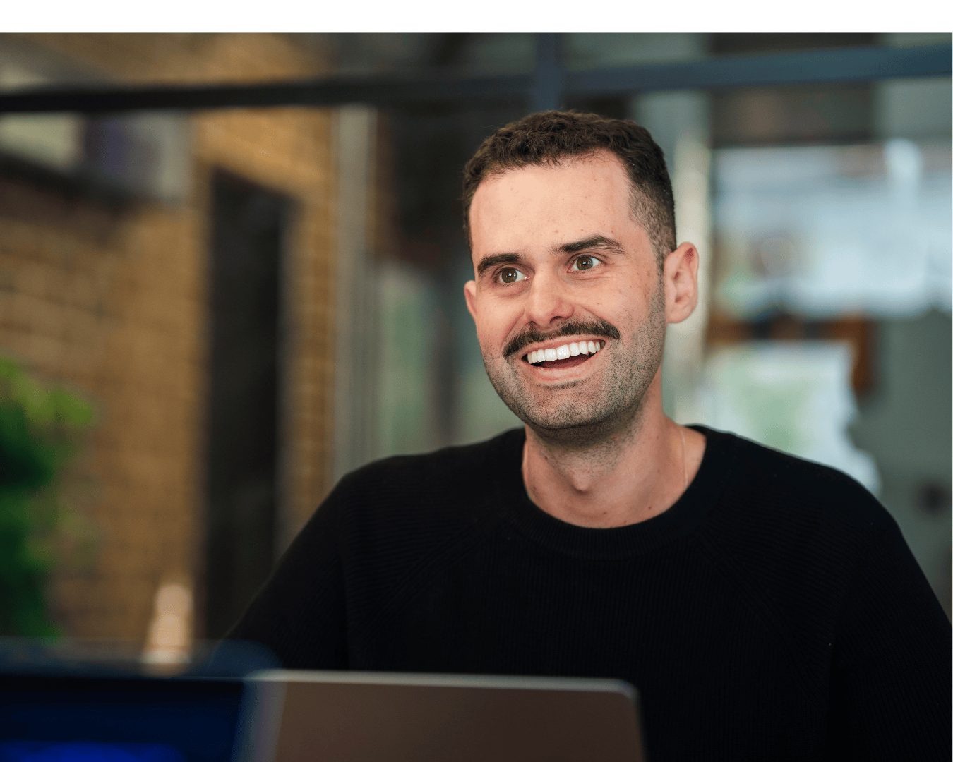 A smiling man with a mustache, wearing a black sweater, sits in a modern office setting. The background features a blurred brick wall and window.