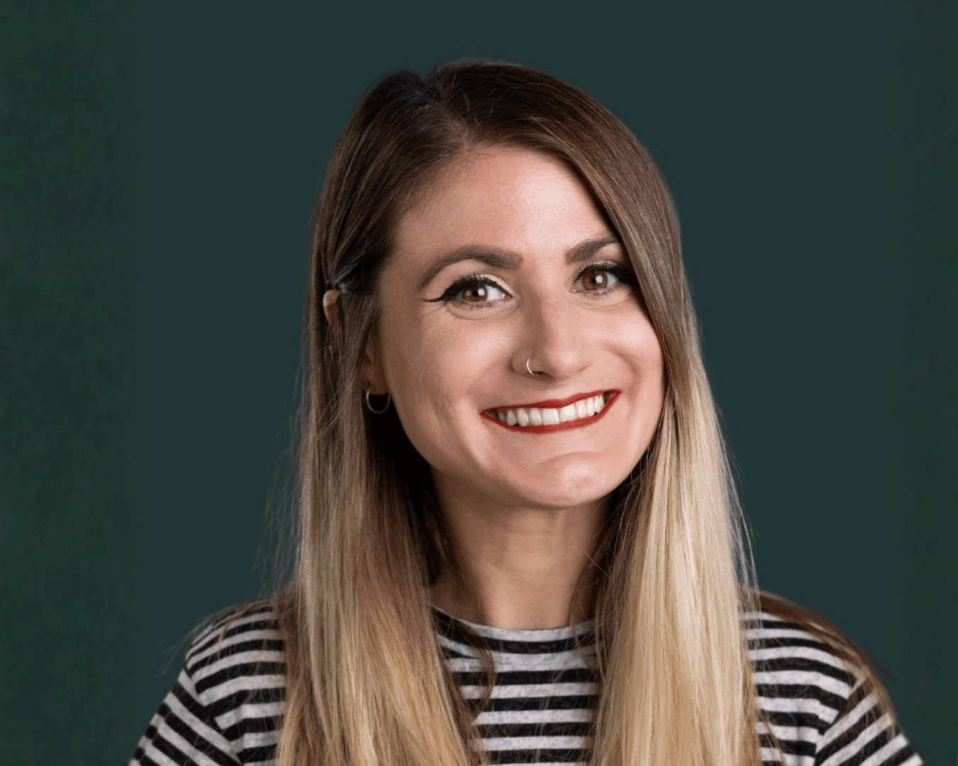 A woman with long, straight hair smiles warmly against a dark teal background. She wears a striped shirt, red lipstick, and hoop earrings