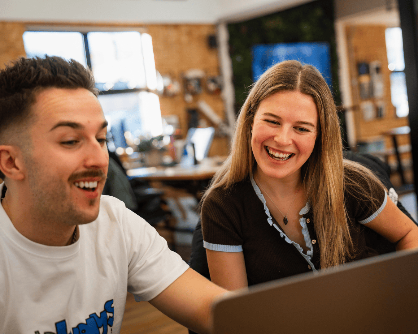Two people smiling and looking at a laptop screen in a cozy office. They appear engaged and happy, with natural light coming through the windows.