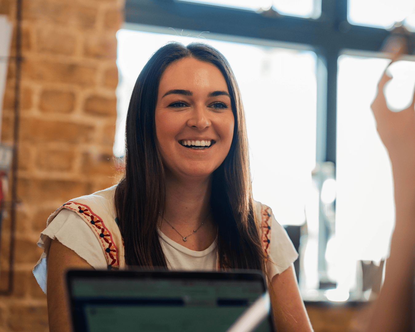 Smiling woman with long hair in casual attire sits in an office setting with exposed brick walls, engaged in conversation with a cheerful expression.