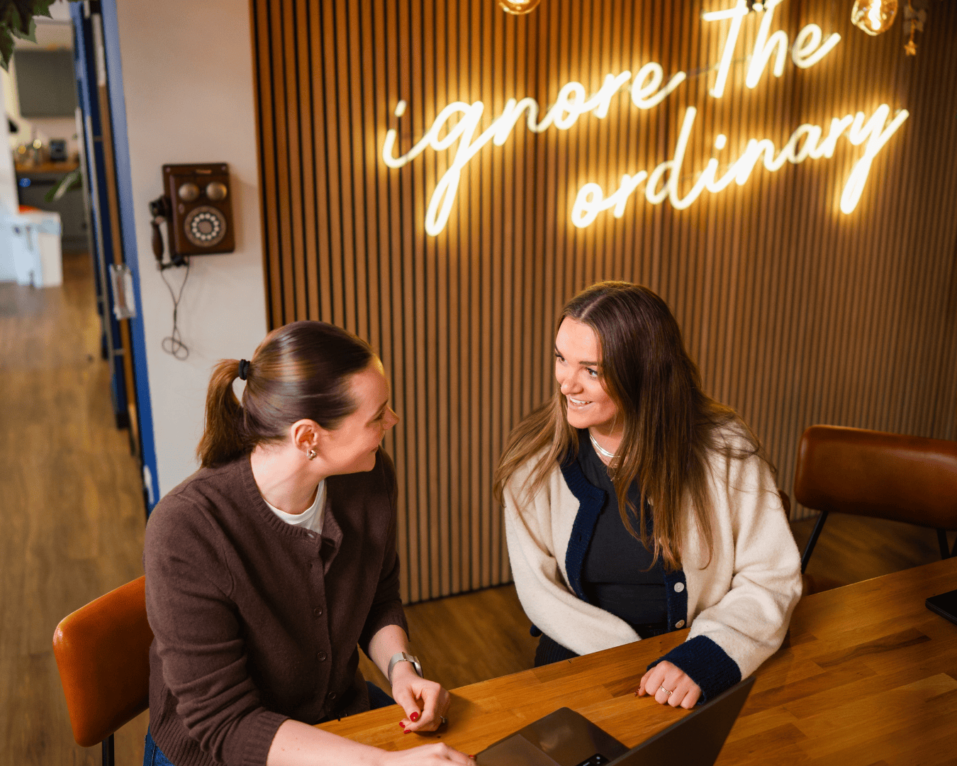 Two women seated at a table, smiling and chatting. A neon sign reading "ignore the ordinary" glows on a wooden wall, creating a warm, inviting atmosphere.