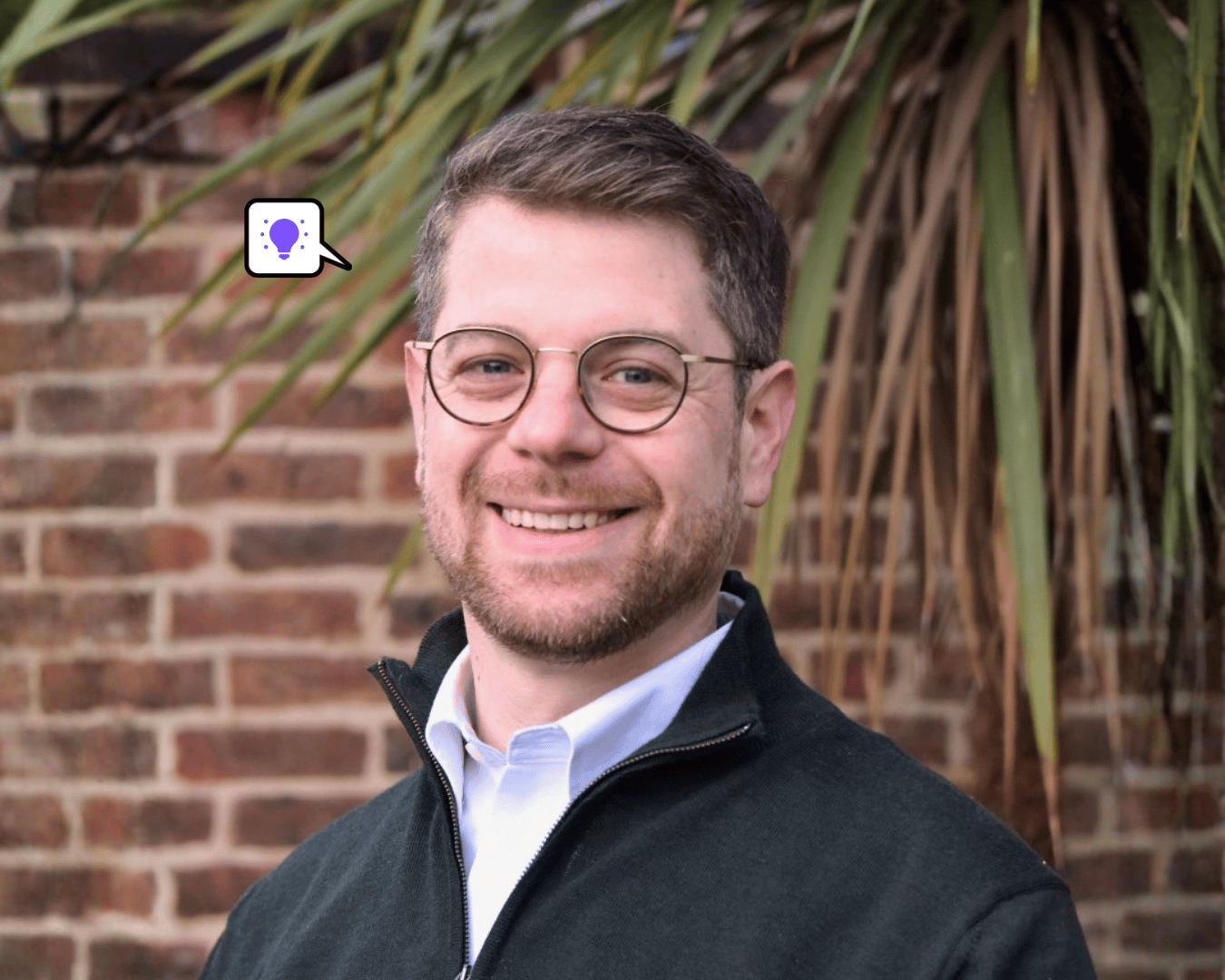 A smiling man with glasses and a beard stands in front of a brick wall and palm leaves.