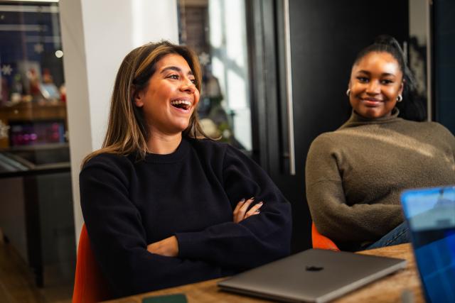 Two women sit at a meeting table in a modern office, smiling during a conversation. The woman on the left appears late 20s–30s with long brown hair, wearing a dark sweater. The woman on the right appears 20s–30s with hair pulled back, wearing an olive knit top.