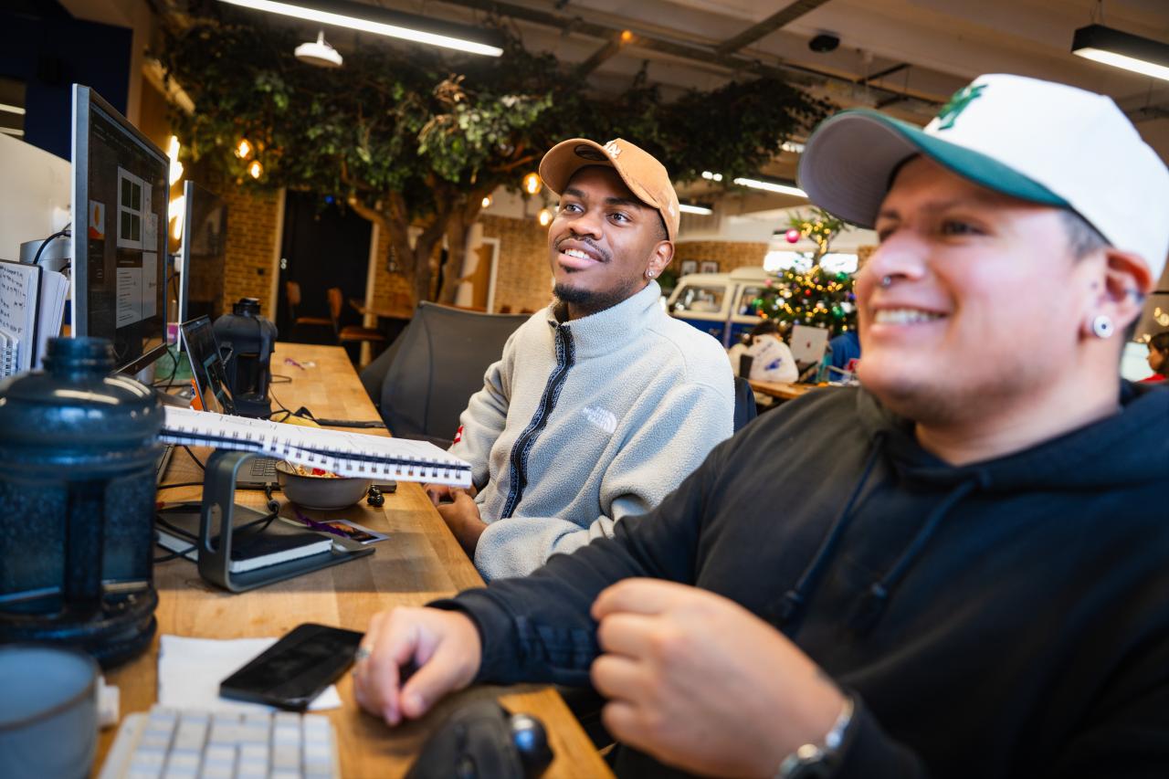Two men sit side by side at desks in a creative office, smiling while looking at a computer screen. Both appear in their 20s–30s. The man on the left wears a beige cap and light fleece jacket; the man on the right wears a green-and-white cap and dark hoodie.