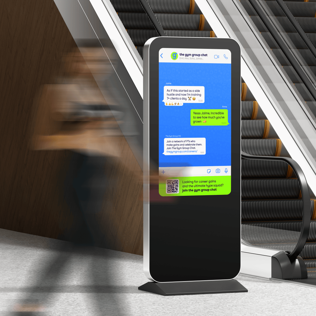 A freestanding digital screen near an escalator displays a group chat interface promoting The Gym Group community. Blurred commuters move past, highlighting high-traffic placement and blending digital recruitment messaging into a fast-paced public environment.
