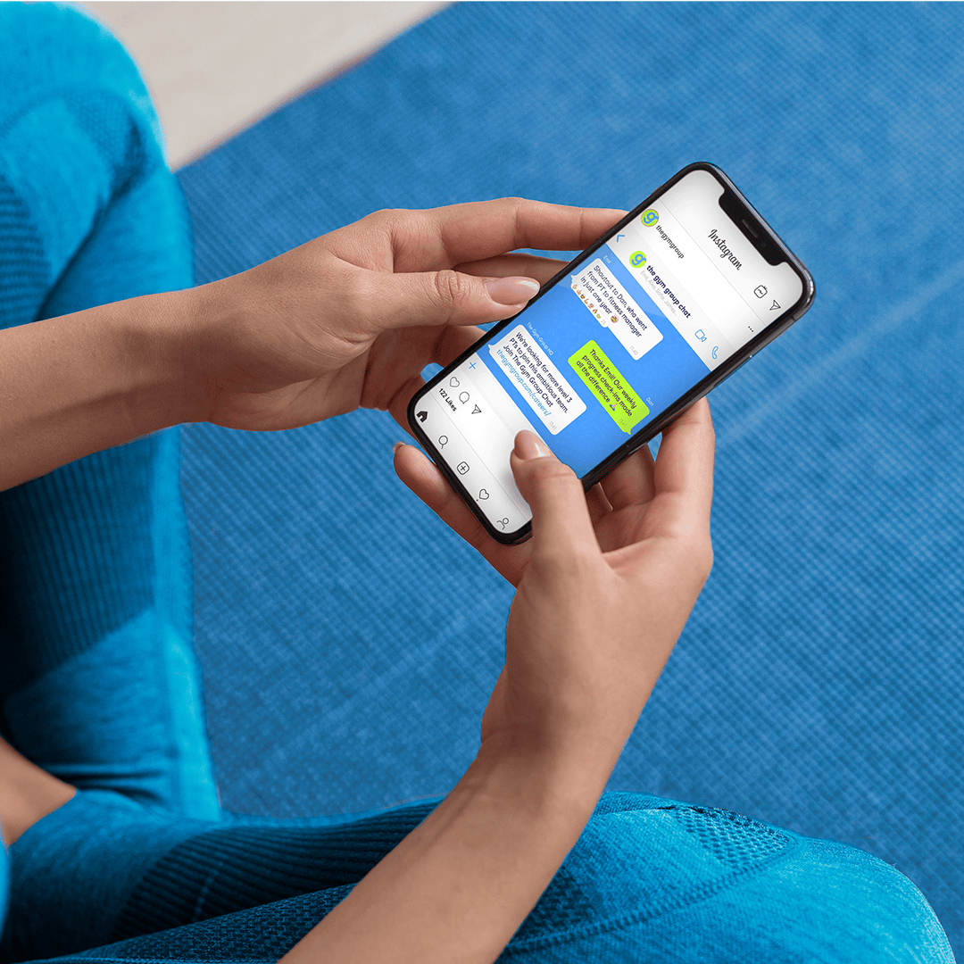 A person in gym clothing sits on a blue exercise mat holding a smartphone. On the screen, an Instagram-style message conversation promotes The Gym Group community, showing supportive messages and career-focused content designed for mobile, social engagement.