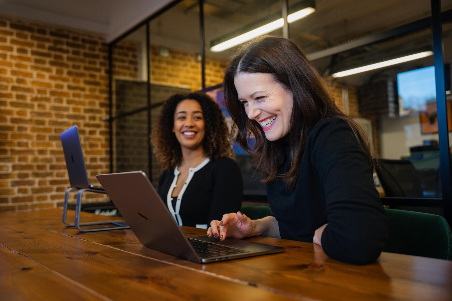 Two colleagues sit at a wooden table in a modern brick office, smiling and laughing while working on laptops. One woman types in the foreground as another looks on, suggesting a friendly, collaborative work moment during a casual meeting together.