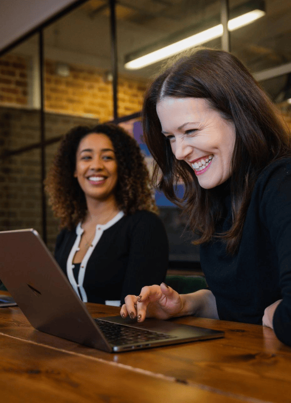 Two colleagues sit at a wooden table in a modern brick office, smiling and laughing while working on laptops. One woman types in the foreground as another looks on, suggesting a friendly, collaborative work moment during a casual meeting together.
