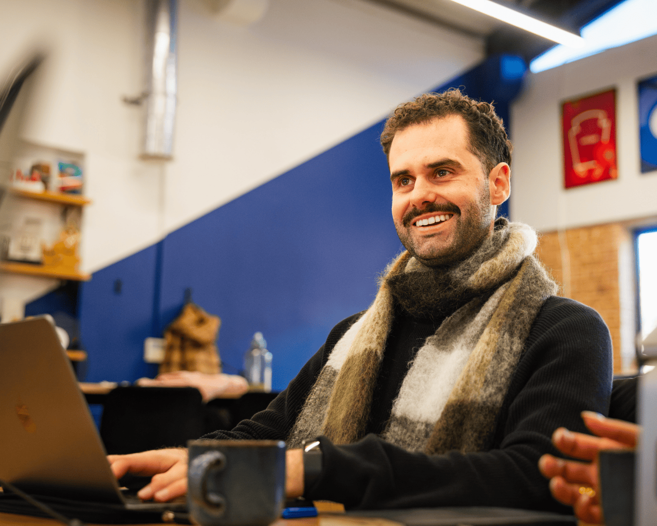 A man with a moustache, wearing a scarf and black sweater, smiles while sitting at a table with a laptop and cup. The setting is a modern office.