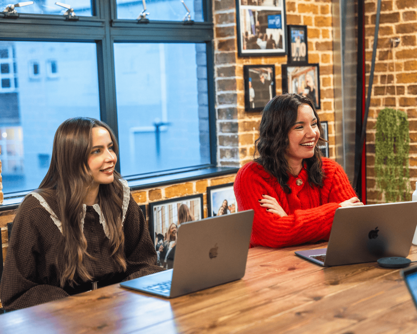 Two women sit at a wooden table in an office with laptops, smiling and engaged. With brick walls and framed photos.