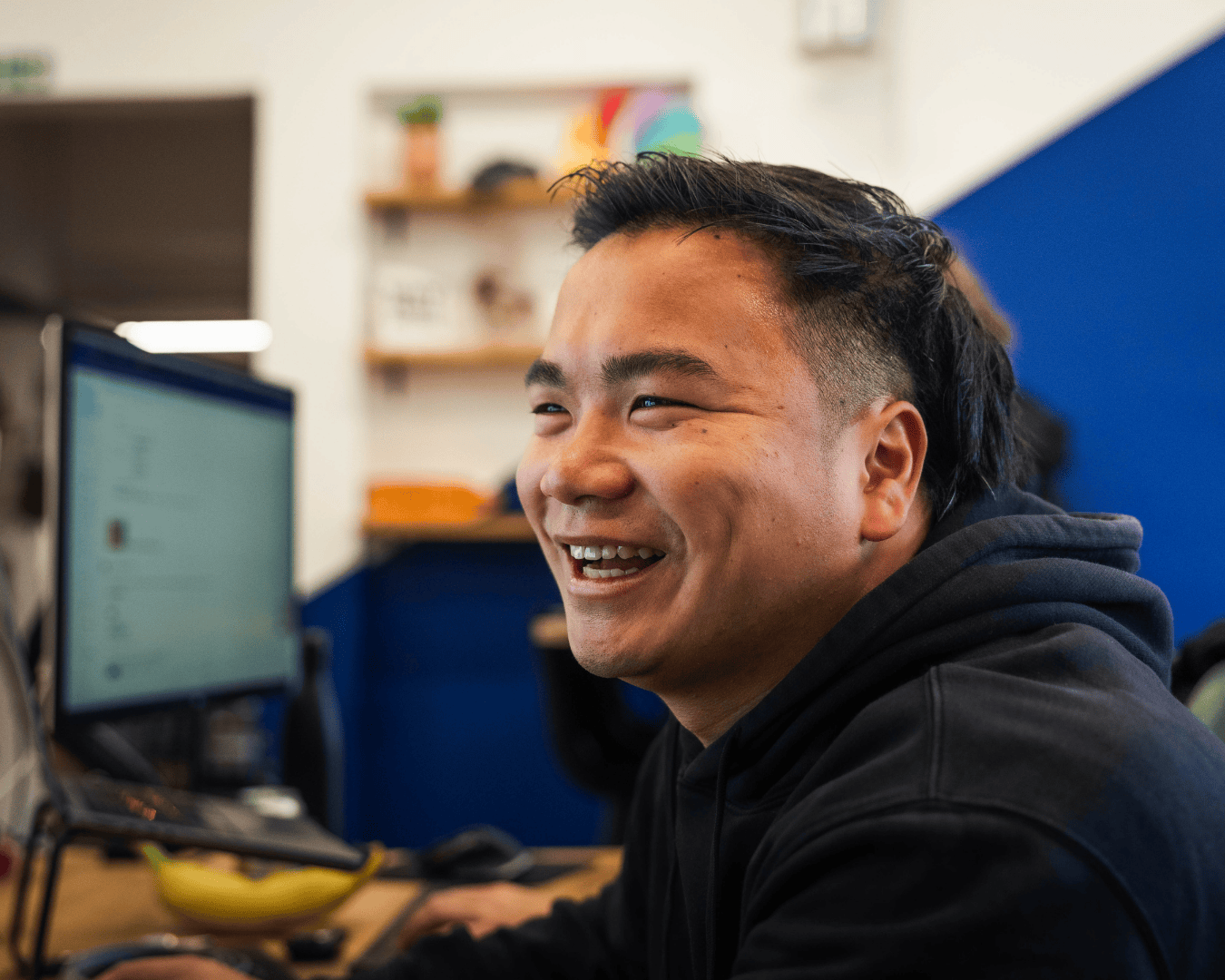 A smiling man in a black hoodie sits at a desk with a computer in an office. The background features blurred shelves and a blue wall.