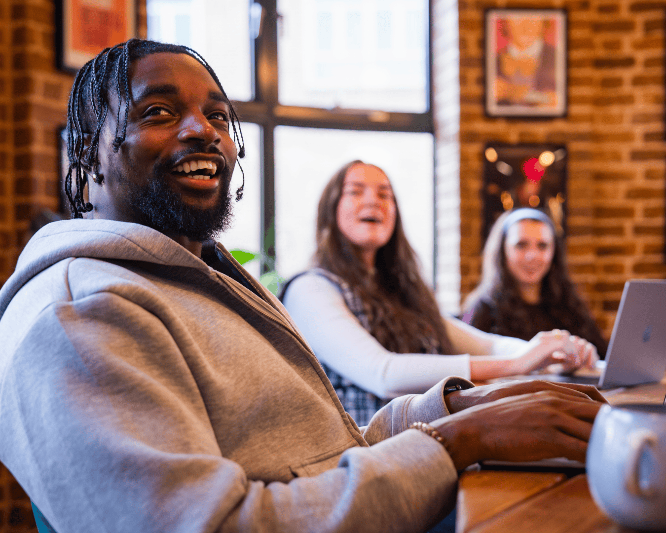 A group of three people sit at a wooden table in a warmly lit room. The foreground male, wearing a grey hoodie. The two females in the background appear engaged. Brick walls and large windows in an office environment