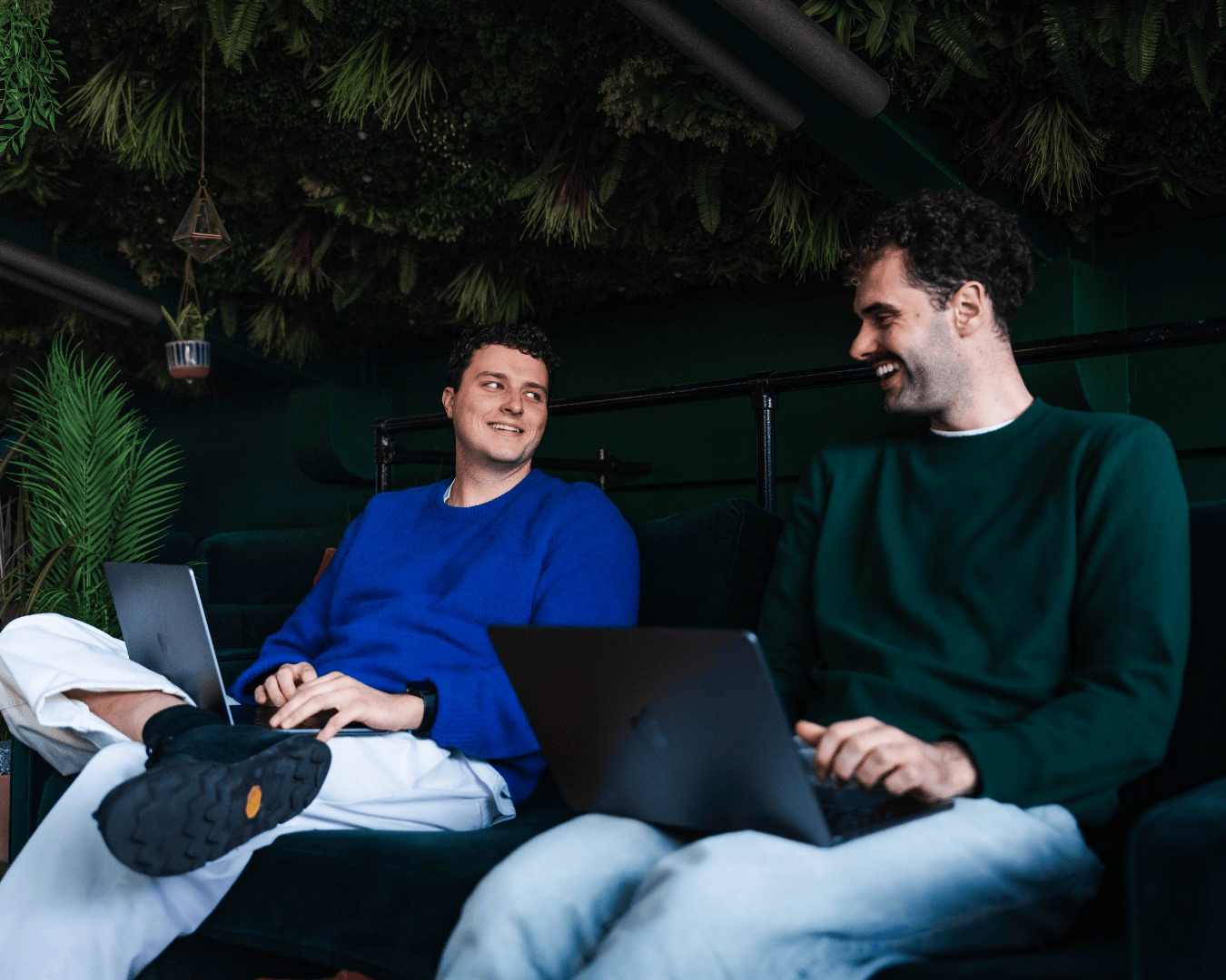 Two people are sitting on a dark green sofa with laptops, smiling at each other. The background features hanging plants