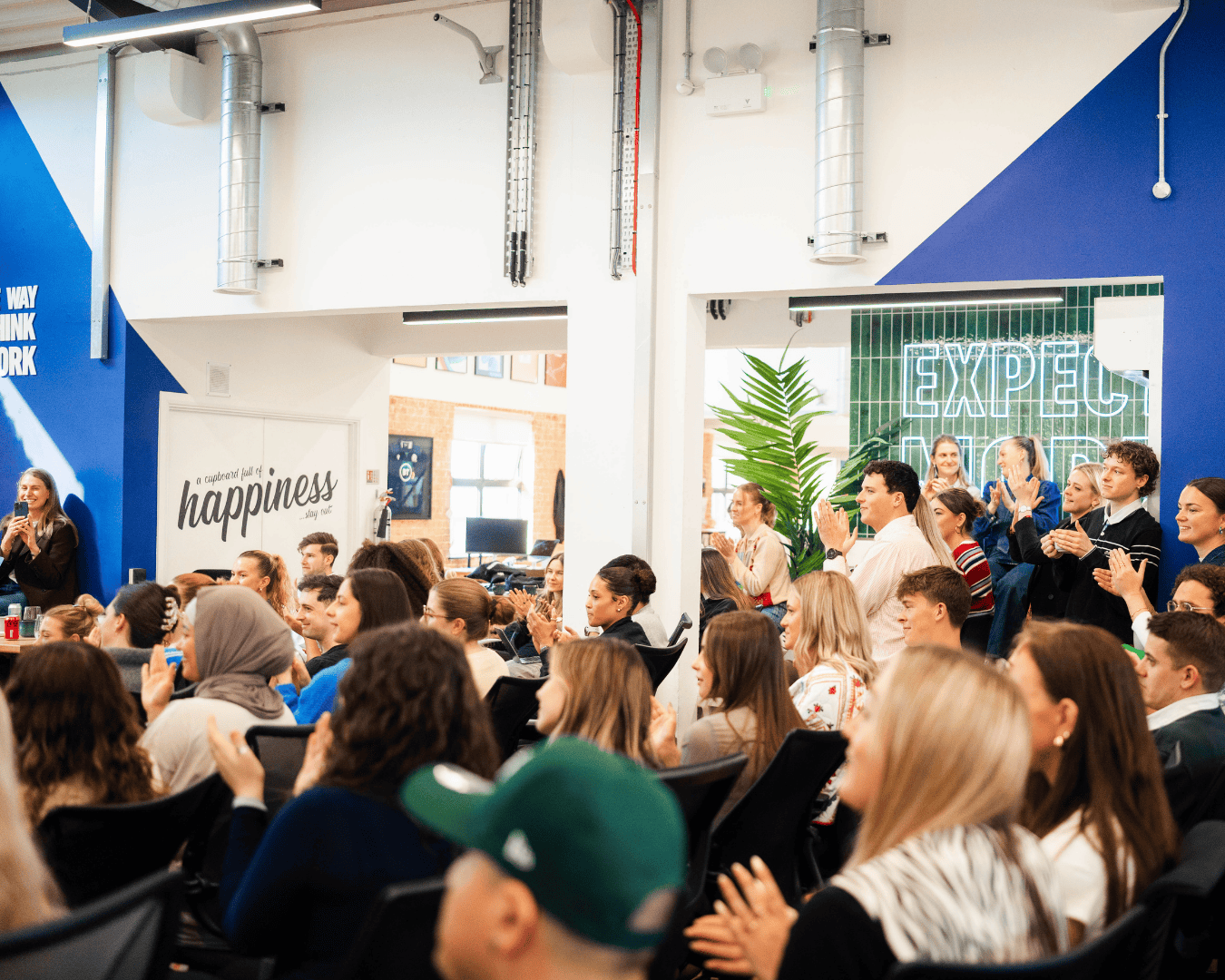 A diverse group of people sit and stand in a bright, modern office space, applauding energetically.