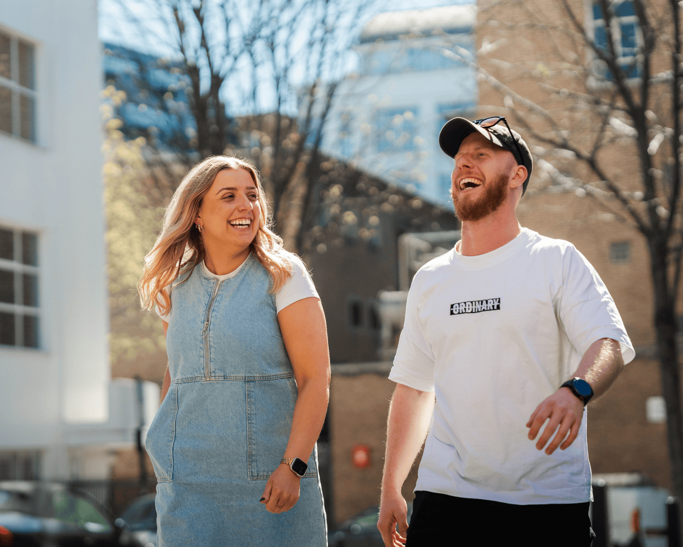A woman in a denim dress and a man in a white T-shirt and cap walk outdoors, laughing. The scene is sunny and relaxed, with buildings in the background.