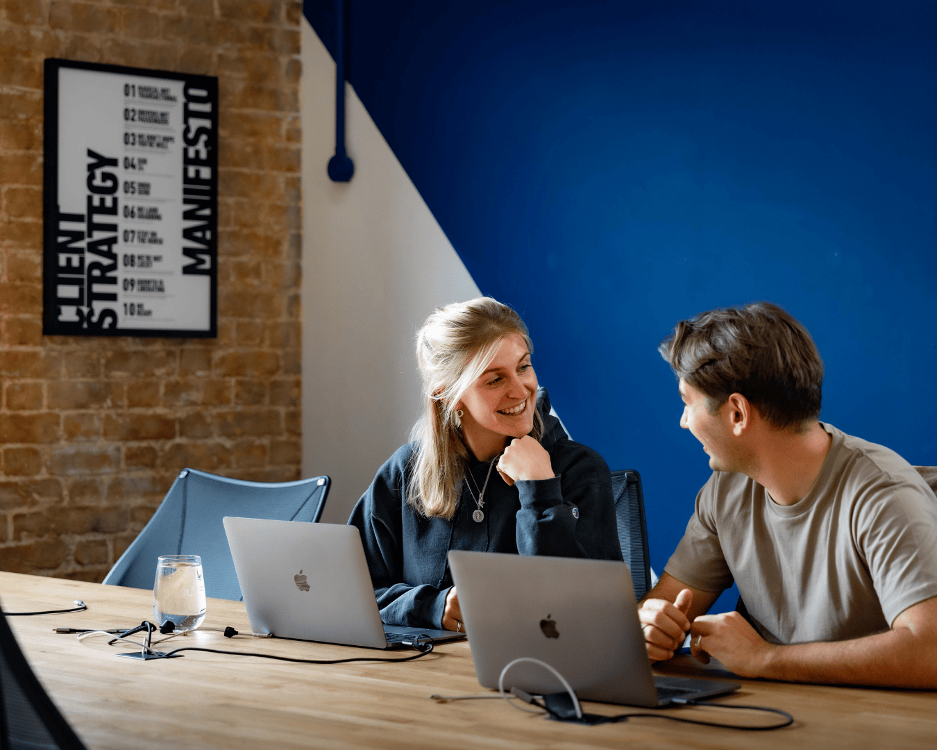 Two people sit at a wooden table with laptops, smiling and talking. A poster with text is on a brick wall.