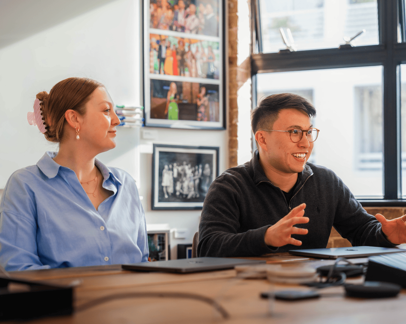 Two people seated at a wooden table, engaged in conversation. A woman in a blue shirt listens attentively while a man in glasses gestures expressively