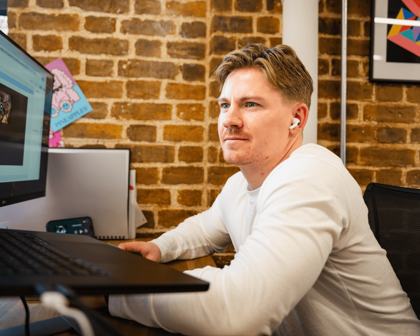 Man with earbuds focused on a computer screen, sitting at a desk against a brick wall. The scene conveys a work atmosphere with concentration.
