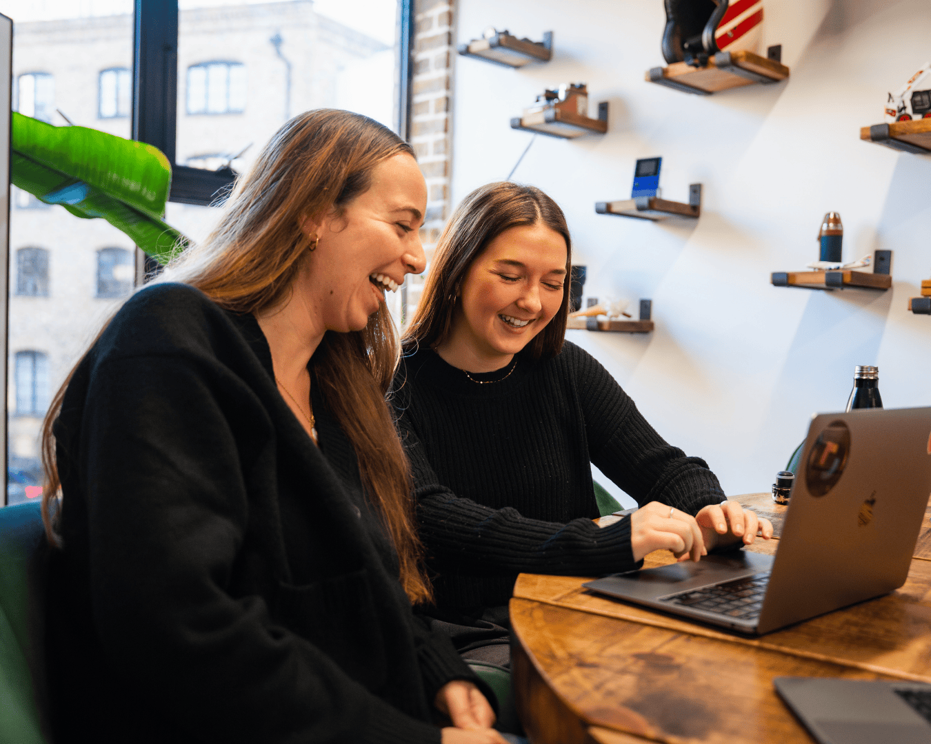 Two women in an office setting are smiling and looking at a laptop screen on a wooden table. Shelves and a window with an urban view are in the background.