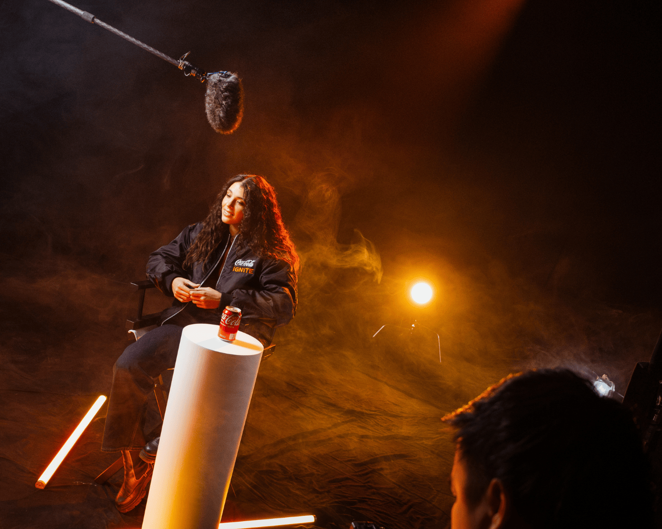 A person with long curly hair sits in a dimly lit studio, wearing a dark jacket. A microphone hangs above, and a can of Coca-Cola is on a white pedestal.