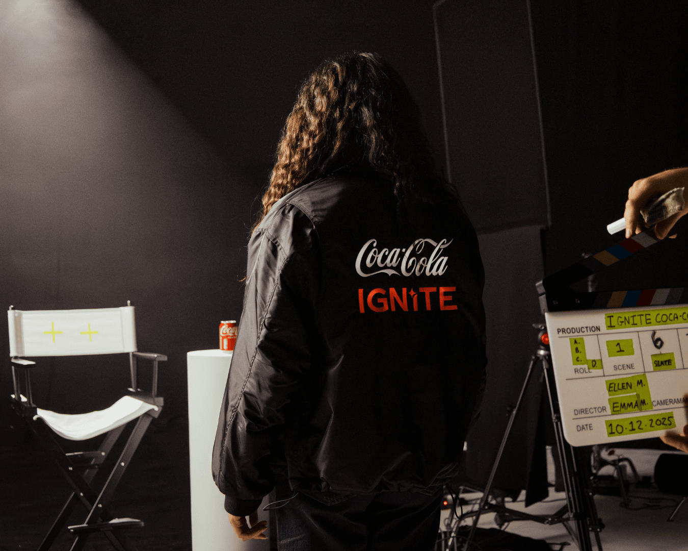 Person with long hair in a "Coca-Cola Ignite" jacket stands in a dimly lit studio. A director's chair, a Coca-Cola can, and a film clapperboard are visible.