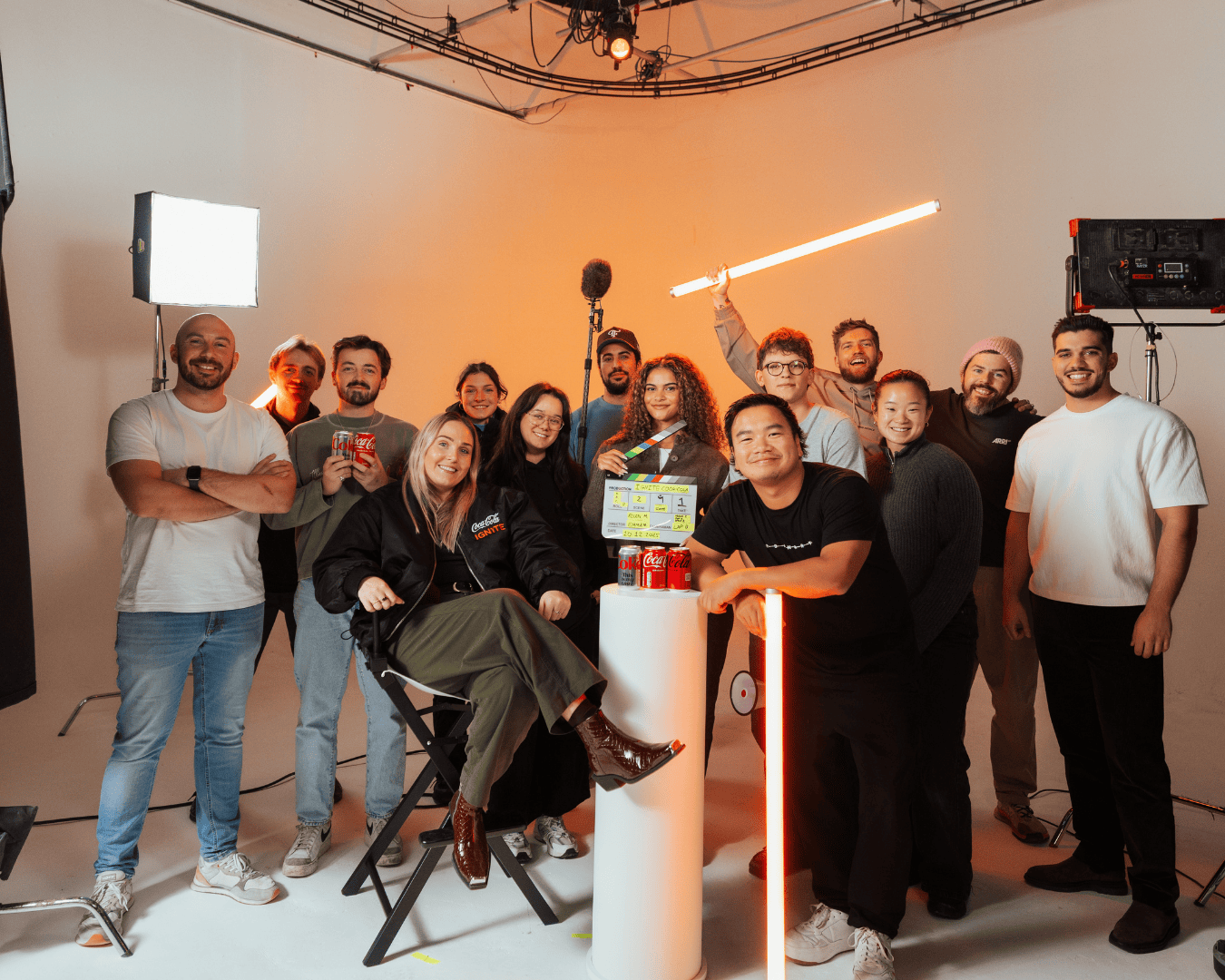 group of 14 people smiling in a brightly lit studio, around a director's chair. They appear cheerful, with some holding drinks and film equipment.