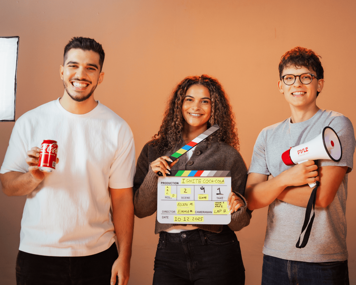Three smiling individuals stand in a studio against a warm background. One holds a Coca-Cola can, another a clapperboard, and the third a megaphone.