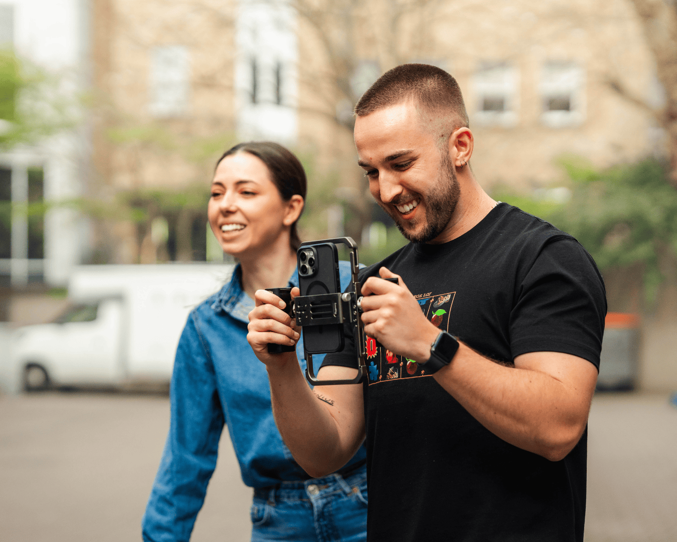 A smiling man with a beard holds a smartphone on a stabilizer, wearing a black T-shirt. A woman in a denim jacket smiles beside him. Outdoor setting.