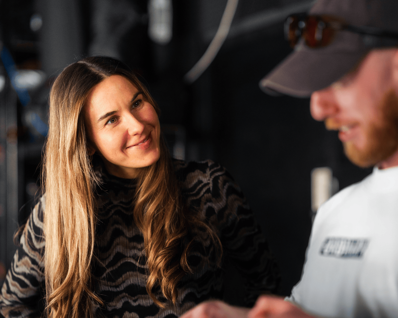 A woman with long hair smiling at a man in a cap, who is looking at a camera.