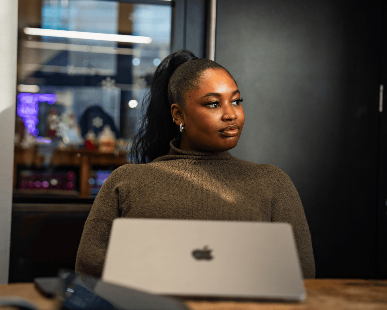 A woman with a ponytail sits thoughtfully at a table with a laptop. She wears a brown sweater. The setting is a modern, well-lit room.