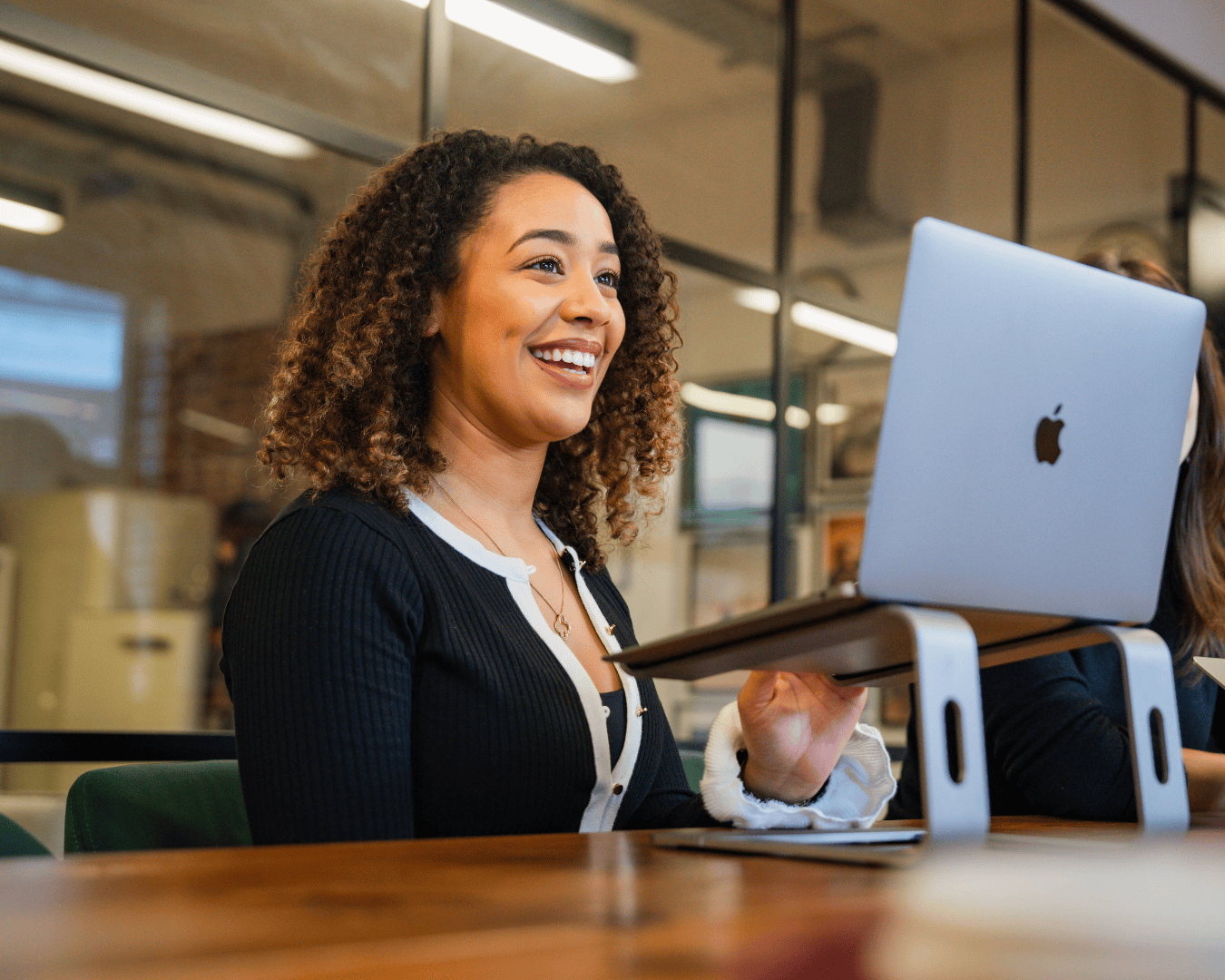 Smiling woman with curly hair working on a laptop at a wooden desk in a modern office. Bright and cheerful atmosphere, with glass walls in the background.