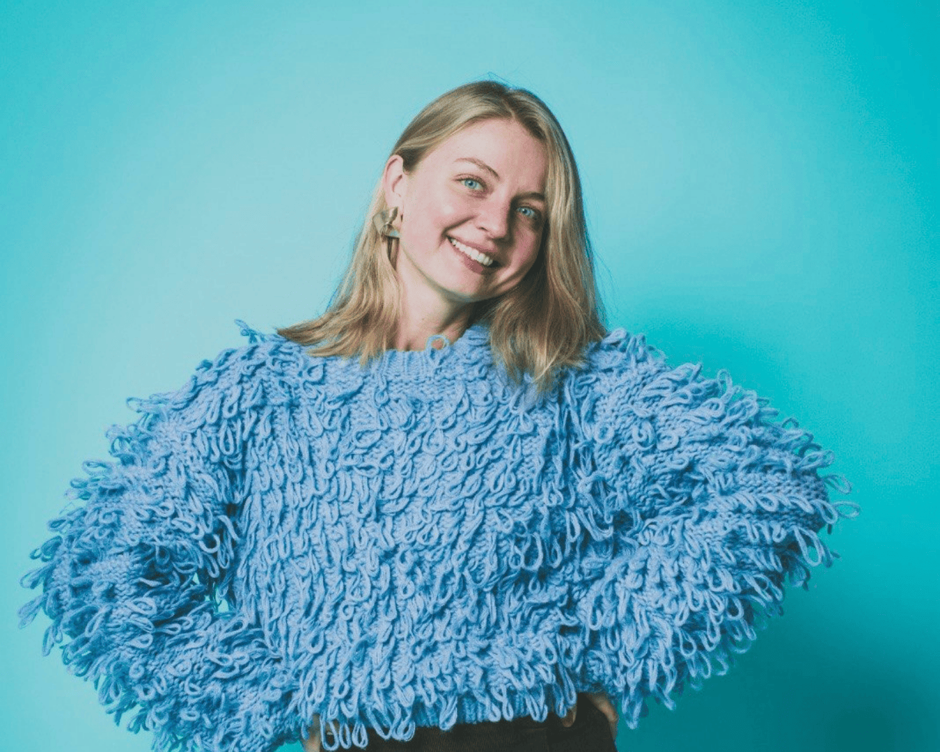 Smiling woman in a textured blue sweater poses with hands on hips against a light blue background.