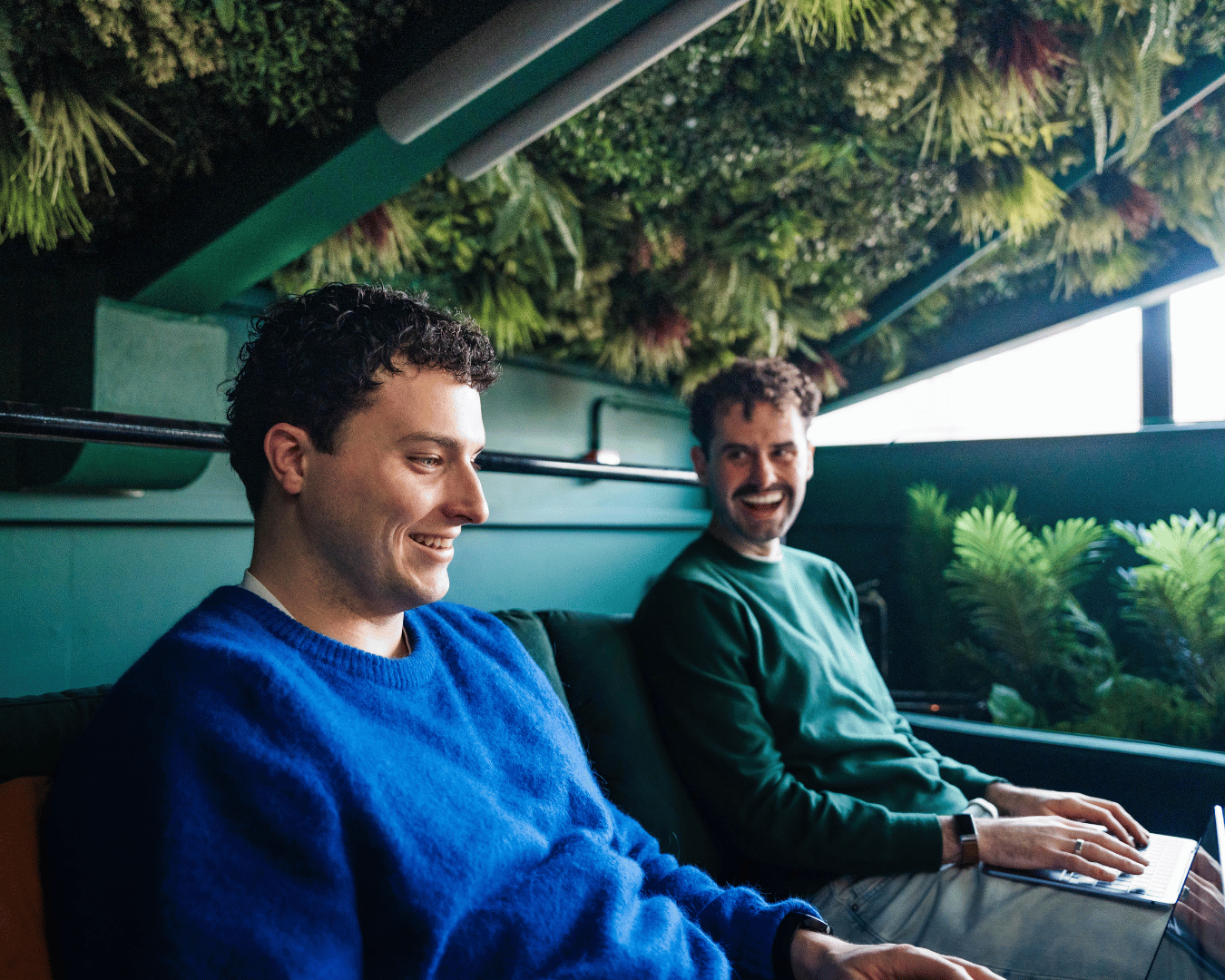 Two men are sitting in a cozy, plant-filled indoor space. They are smiling and appear relaxed, with one using a laptop. A ceiling of greenery adds a natural vibe.