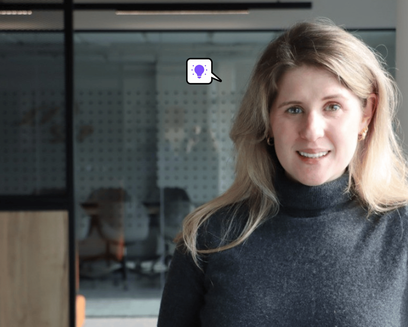 Woman with long hair in a gray turtleneck smiles slightly, standing in a modern office with glass walls. A thought bubble with a light bulb icon appears.