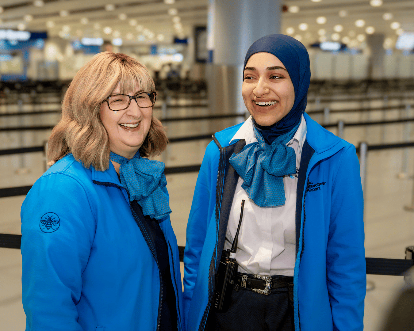 Two smiling employees in blue uniforms stand in an airport terminal. They wear matching scarves and blue jackets, creating a welcoming atmosphere.